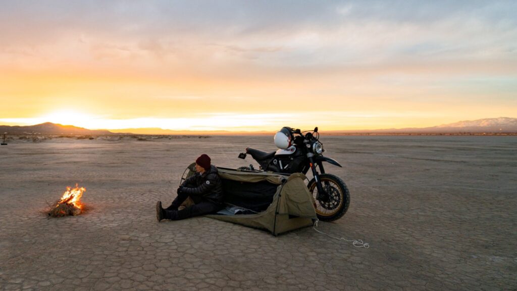 Man on a motorcycle camping trip with fire burning and sunset in the distance